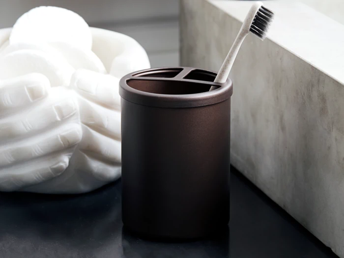 En sort tandbørsteholder med en tandbørste indeni står på en mørk disk ved siden af en hvid skulptur af hænder, der holder runde genstande.