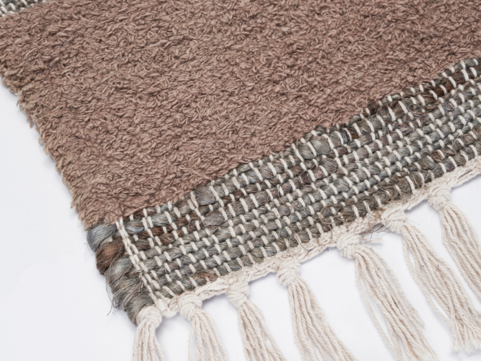 Et nærbillede af et vævet brunt og beige tekstil med frynsede kvaster langs kanten, vist på en hvid baggrund.