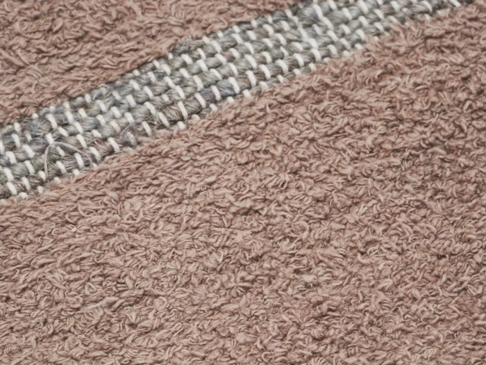 Nærbillede af et struktureret brunt stof med en vandret stribe af tætvævede, lysere og mørkere tråde nær toppen.