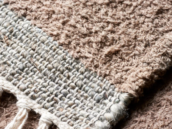 Nærbillede af et struktureret, vævet brunt stoftæppe med synlige fibre og en flettet kant, der viser detaljeret vævning og løse tråde.