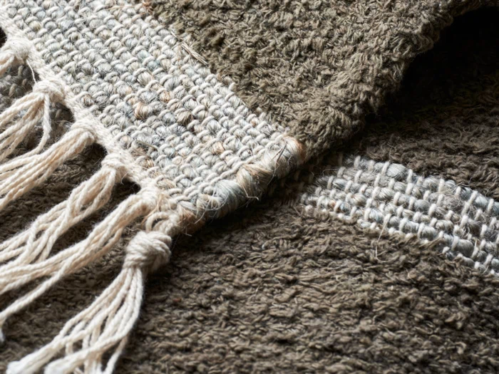 Nærbillede af et vævet stof med tykke, strukturerede fibre i jordfarver og med knyttede frynser på den ene kant.