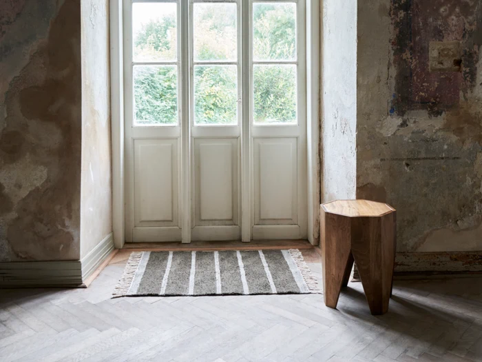 Et rustikt rum med slidte vægge, en træskammel, et stribet tæppe og store glasdøre med hvid ramme, der lukker naturligt lys ind.