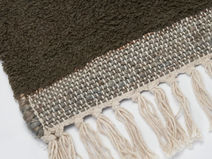 Nærbillede af et vævet tekstil med en mørkebrun struktureret del, et lysere vævet område og cremefarvede frynsede kvaster langs kanten.