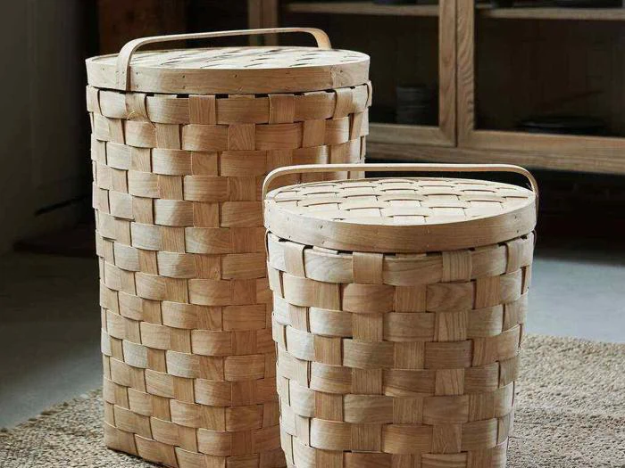 To flettede, lyse kurve med låg og håndtag er placeret på et tekstureret tæppe indendørs.