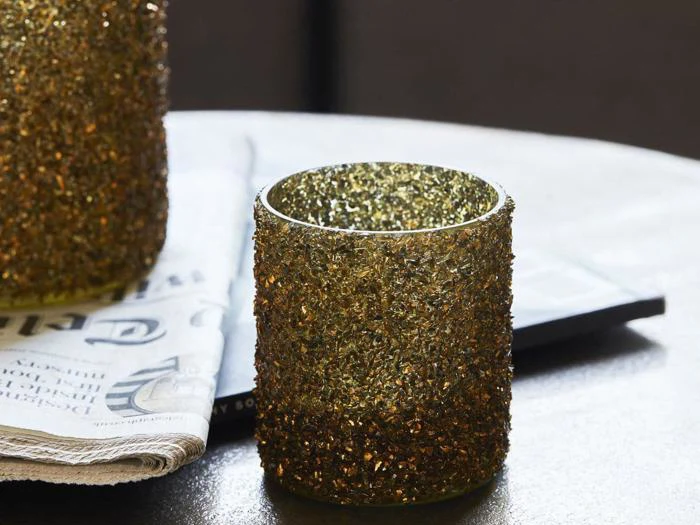 En guldfarvet, struktureret glaslysestage står på et bord ved siden af en sammenfoldet avis og en anden lignende lysestage i baggrunden.