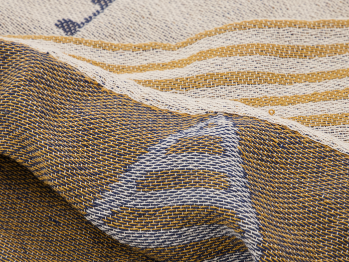 Nærbillede af et struktureret vævet stof med cremefarvede, blå og guldfarvede tråde, der danner striber og trekantede mønstre.