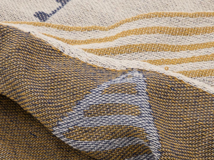 Nærbillede af et struktureret vævet stof med cremefarvede, blå og guldfarvede tråde, der danner striber og trekantede mønstre.