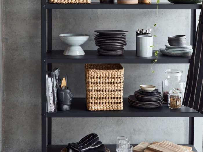 Sort reol med stablede keramikskåle og -tallerkener, en flettet kurv, glaskrukker, redskaber i en holder og en hængeplante mod en betonvæg.