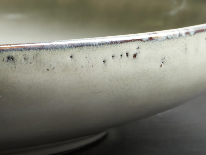 Nærbillede af kanten på en keramikskål med en glat, lys glasur og små mørke pletter på en mørk baggrund.