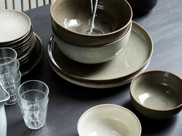 Et sæt keramiktallerkener, skåle og klare drikkeglas står pænt på en mørk bordplade, og i en af skålene ligger en metalske.
