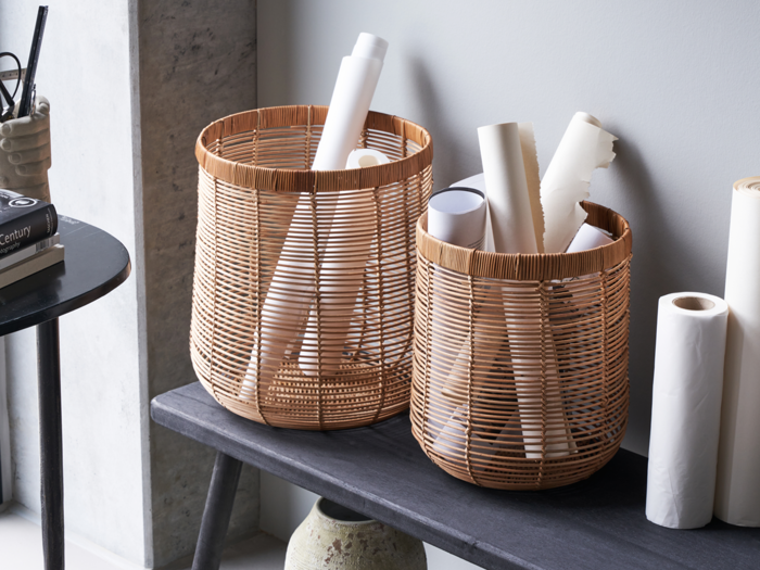 To flettede kurve på en bænk indeholder sammenrullede hvide papirer; et sort bord med bøger og en kop med kuglepenne står ved siden af en lysegrå væg.