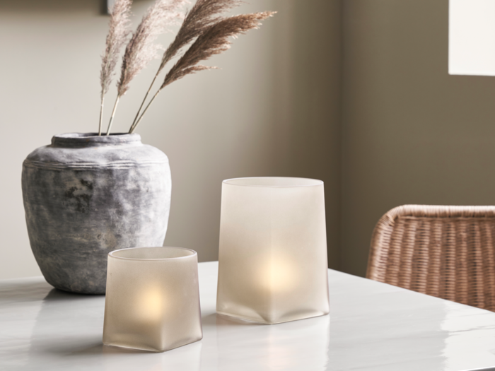 Et marmorbord med to lysestager i matteret glas, en rustik grå vase med tørret pampasgræs og en vævet stol i baggrunden.