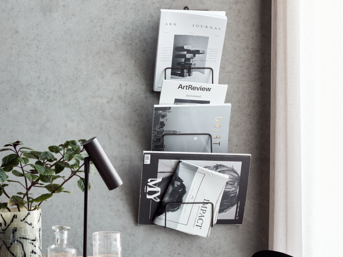 En væghængt magasinholder med flere magasiner og tidsskrifter ved siden af et skrivebord med en sort lampe, en plante og et glas.