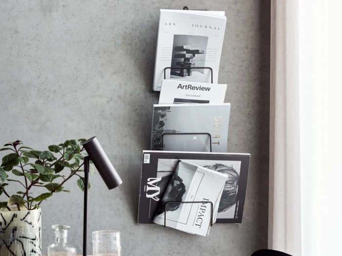 En væghængt magasinholder med flere magasiner og tidsskrifter ved siden af et skrivebord med en sort lampe, en plante og et glas.