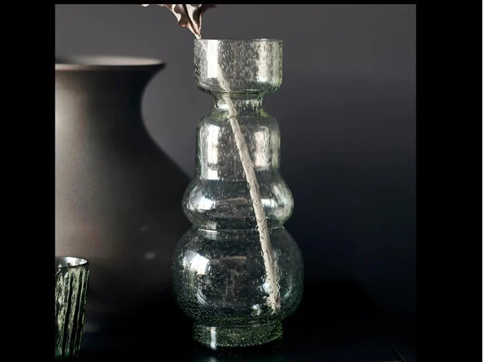 En klar, struktureret glasvase med smal hals og afrundet krop står på en mørk overflade mod en mørk baggrund. Et tørret blad er placeret inde i vasen.