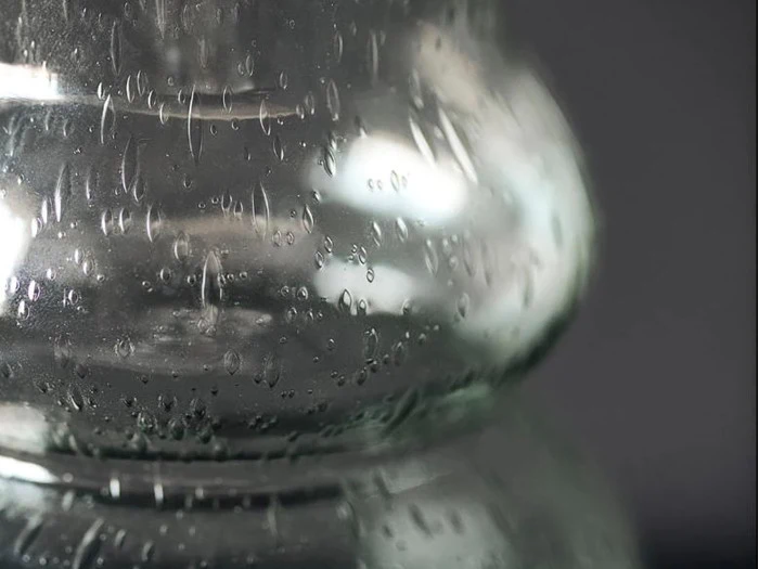 Nærbillede af en klar glasvase med vanddråber og kondens på den buede overflade mod en mørk baggrund.