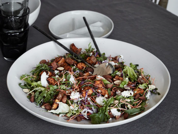 En stor hvid tallerken med en blandet salat af grønt, ristede søde kartofler, mandler og osteklatter på en mørk dug, med serveringsredskaber og tomme tallerkener i nærheden.
