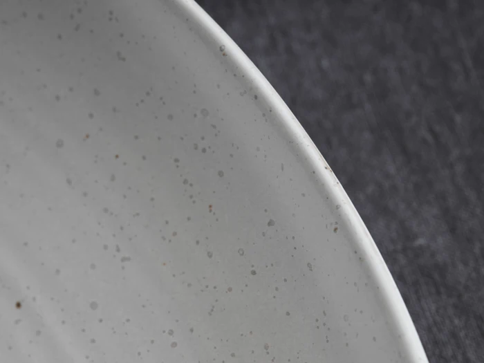 Nærbillede af kanten af en hvid keramiktallerken med små brune pletter på en mørk, struktureret baggrund.