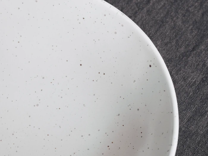 Nærbillede af en hvid keramiktallerken med små brune pletter, placeret på en mørk stofbaggrund.