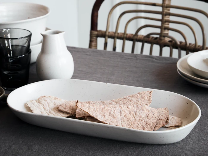 Et hvidt keramikfad med stykker af knækbrød står på en grå dug med glas, en lille hvid kande, tallerkener og en rattan-stol i baggrunden.