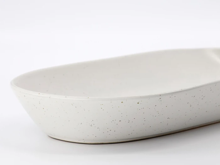 Et nærbillede af et hvidt, ovalt keramikfad med et spættet mønster, placeret på en almindelig hvid baggrund.