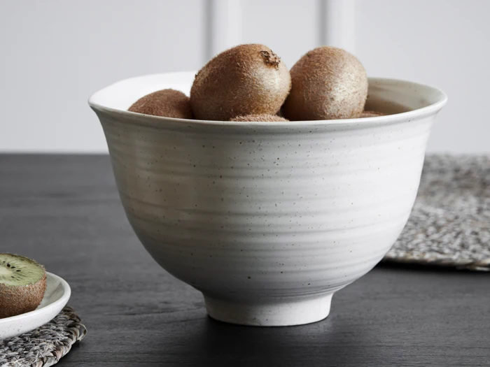 En hvid keramikskål fyldt med hele kiwier står på et mørkt bord med en kiwi i skiver på en måtte ved siden af.