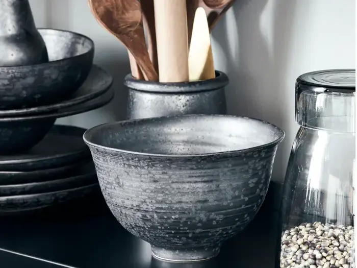 En keramisk skål med en spættet grå finish står på en sort bordplade ved siden af stablede tallerkener, træredskaber i en holder og en glaskrukke fyldt med bønner eller frø.