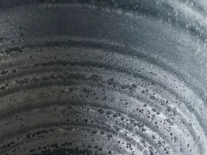 Nærbillede af en metallisk overflade med cirkulære mønstre og små mørke pletter, der ligner ringe eller riller med et struktureret, forvitret udseende.