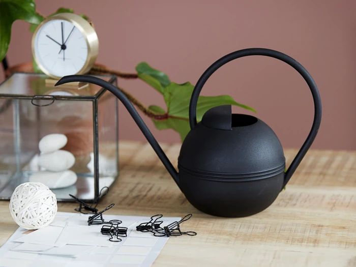 En moderne sort vandkande på et træbord med et ur, en glaskasse med hvide sten, clips, en snor og en grøn plante i baggrunden.