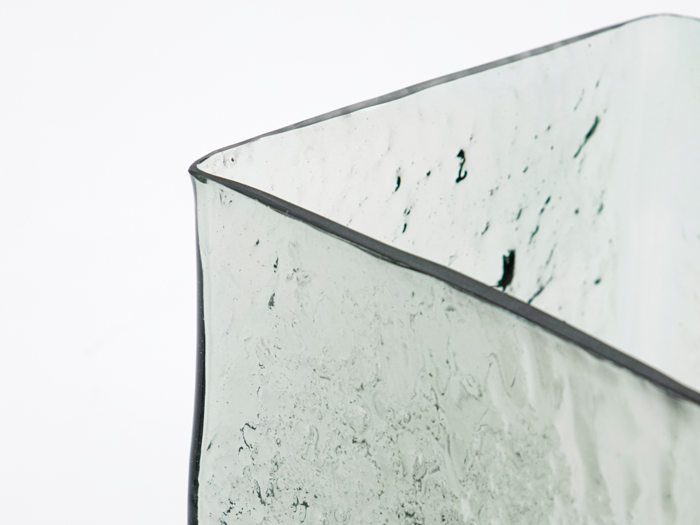 Nærbillede af en gennemsigtig, struktureret glasbeholder med bølgede, ujævne kanter mod en almindelig hvid baggrund.