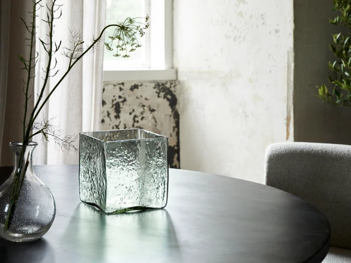 En firkantet struktureret glasvase og en rund glasvase med en tynd gren står på et mørkt rundt bord i et blødt oplyst rum med et vindue og neutral indretning.