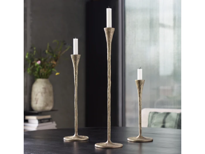 Tre metalliske lysestager i forskellige højder med hvide stearinlys placeret på et mørkt bord. En vase med blomster og stablede bøger er i den slørede baggrund.