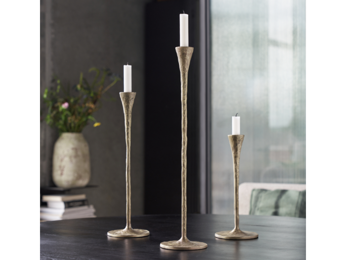 Tre metalliske lysestager i forskellige højder med hvide stearinlys placeret på et mørkt bord. En vase med blomster og stablede bøger er i den slørede baggrund.