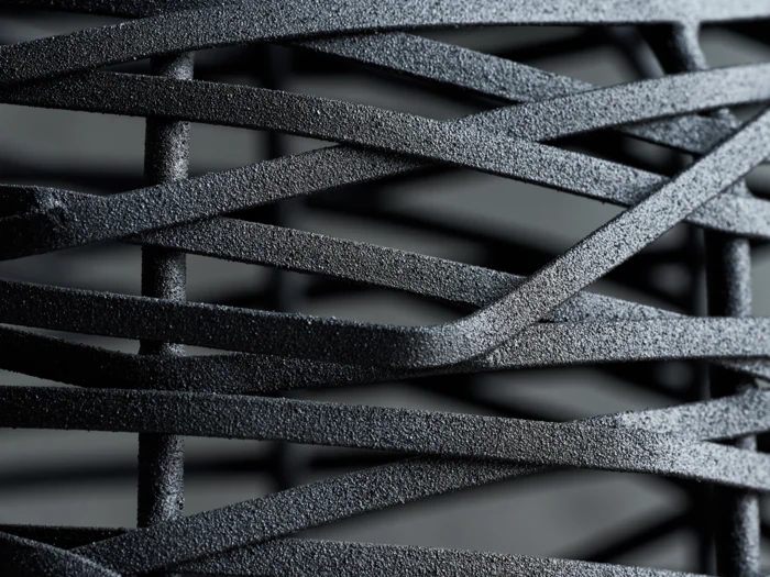 Nærbillede af overlappende, teksturerede sorte metal- eller plastikbånd arrangeret i et krydsende mønster, der skaber et abstrakt, geometrisk design.