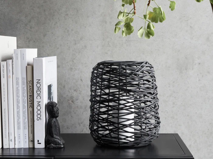 En sort vævet lampe, bøger og en lille buddhastatue er arrangeret på et sort bord mod en grå betonvæg med grønne blade hængende over.