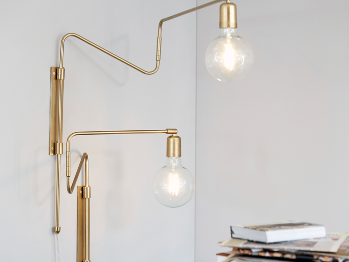 En moderne væglampe i messing med synlige pærer og justerbare arme er monteret på en hvid væg ved siden af en stak bøger og magasiner.