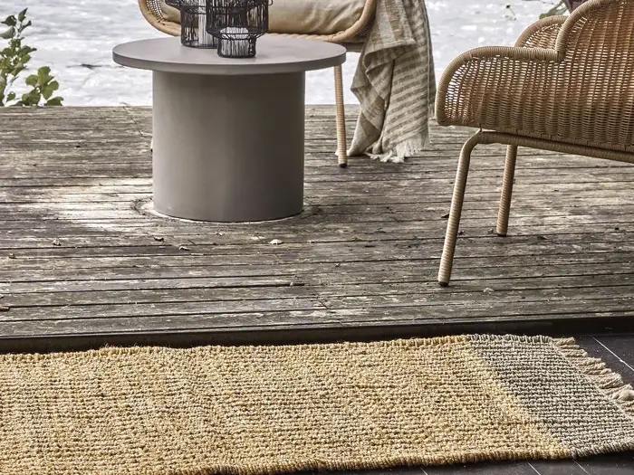 Et vævet tæppe ligger på en træterrasse med et rundt bord, kurvestole og et stribet tæppe, der er draperet over en stol nær et vandløb.