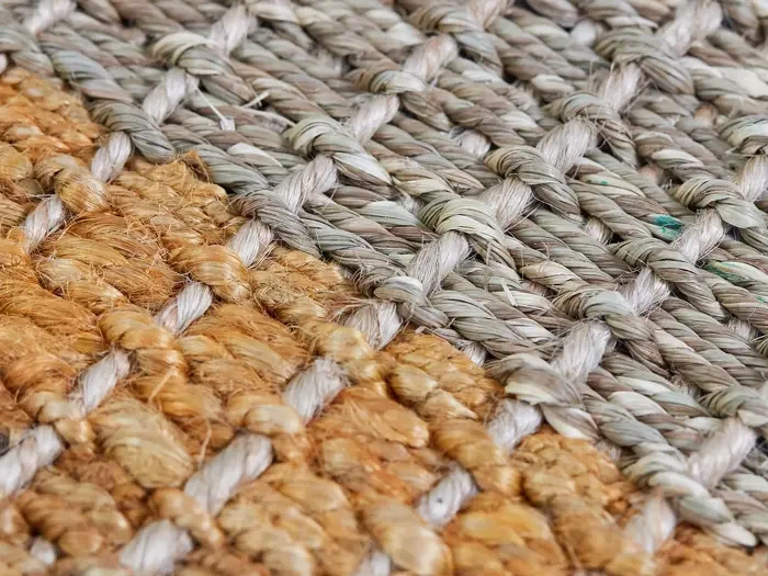 Nærbillede af et vævet tæppe med tykke, strukturerede fibre i to farver: naturbrun og lysegrå, arrangeret i et kurvefletningsmønster.