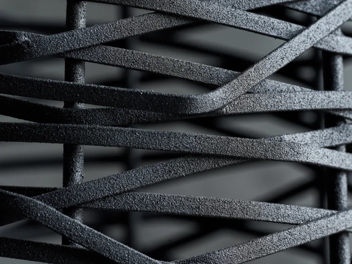 Nærbillede af sorte, strukturerede metal- eller plastikstrimler på kryds og tværs, som skaber et mønster med krydsende linjer og skygger.
