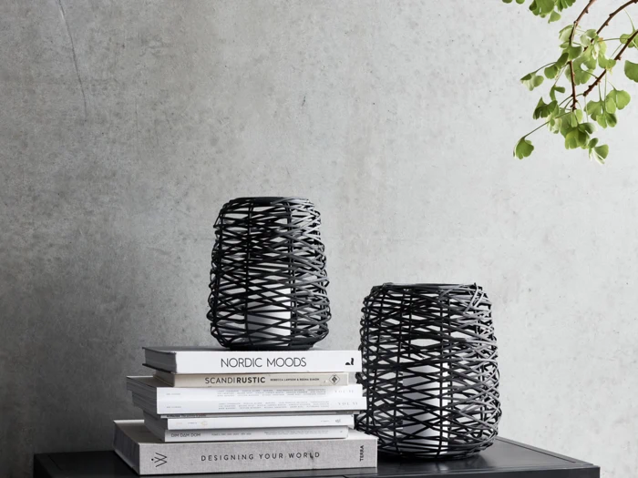 To sorte flettede vaser står på en stak bøger op ad en grå betonvæg med grønne blade hængende fra øverste højre hjørne.