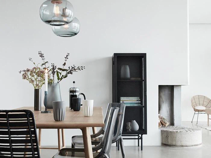 Moderne spisestue med træbord, klare stole, vaser med blomster, pendler, sort skab og minimalistisk pejs mod hvide vægge.