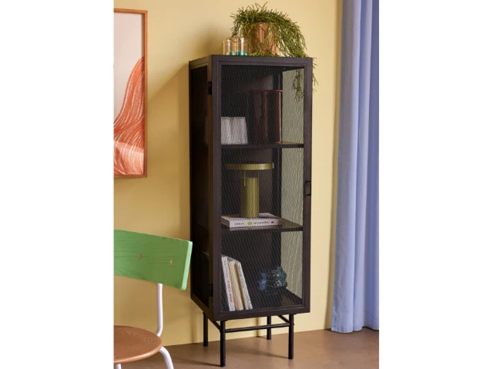 Et højt sort metalskab med netdør står op ad en gul væg med bøger, indretning og en lampe, mens en grøn stol og indrammet kunst står i nærheden.