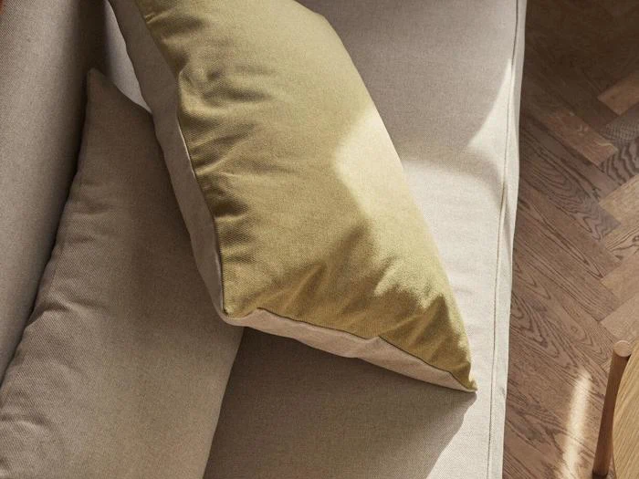 En beige sofa med to puder, en beige og en lys olivengrøn, står på et træparketgulv i naturligt sollys.