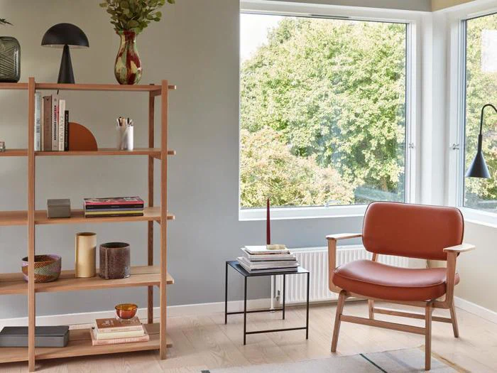 En moderne stue med læderstol, sidebord med bøger, bogreol i træ og store vinduer med udsigt til grønne træer.