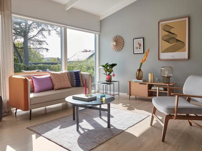 Moderne stue med beige sofa, farverige puder, træmøbler, stort vindue med udsigt til haven og moderne vægkunst. Sollyset lyser op i det hyggelige, stilfulde rum.