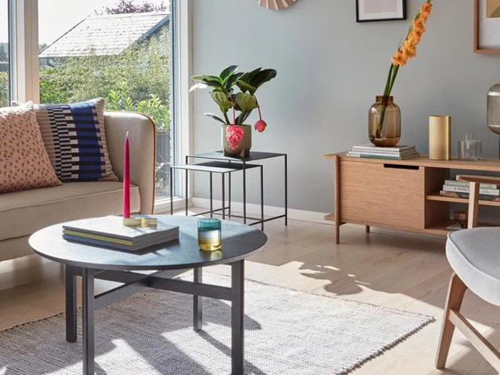 En moderne stue med en lys sofa, mønstrede puder, et rundt sofabord, et tæppe, potteplanter og sollys, der strømmer ind gennem et stort vindue.