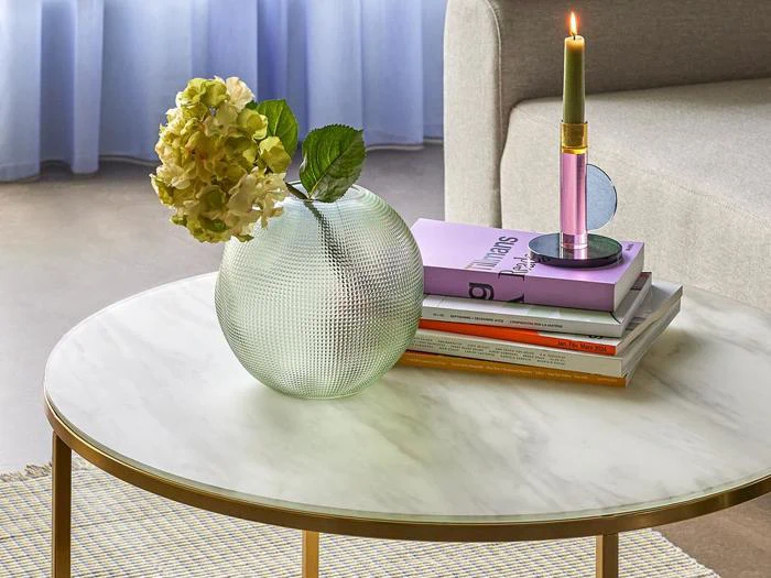 Et rundt sofabord i marmor med en tekstureret glasvase med en hortensia, en stak bøger og et tændt tofarvet stearinlys i en holder.