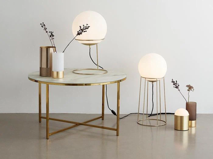 På et moderne rundt bord med guldben står to minimalistiske vaser og en glødende globelampe; på gulvet ved siden af står yderligere to globelamper og en vase.