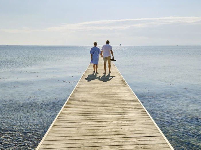 To mennesker går side om side på en træmole, der strækker sig ud over roligt havvand, på vej mod horisonten under en klar himmel.