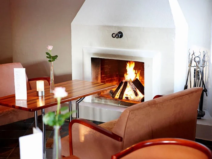 Hyggeligt opholdsområde med brune stole og et træbord ved en tændt pejs; en enkelt rose i en vase og et stearinlys står på bordet.